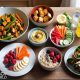 kitchen table with a colorful layout of healthy diet foods — include a bowl of mixed vegetable stir-fry, grilled tofu, a plate of hummus with carrot and cucumber sticks, fresh fruit salad, a bowl of oatmeal topped with banana and flaxseeds, a bottle of infused lemon water, and a whole grain wrap filled with lean protein and greens.